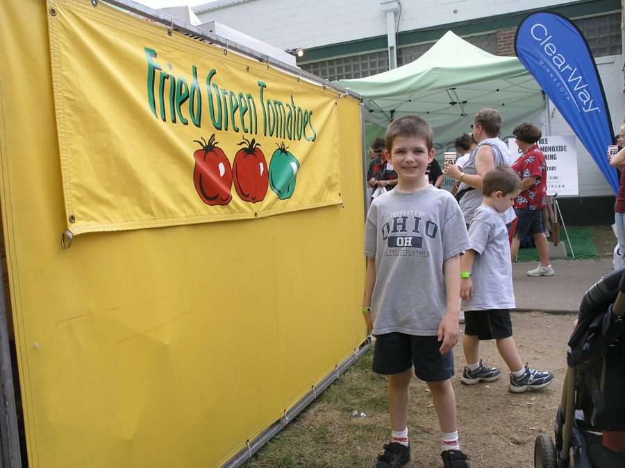 A boy posing proudly beside the stand's yellow Fried Green Tomatoes banner with its three-tomato logo